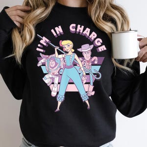 Może przedstawiać: Czarna bluza z nadrukiem graficznym postaci z franczyzy Toy Story. Tekst "I'm in charge" jest nadrukowany na różowo-fioletowym tle.