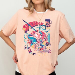 K&ouml;nnte beinhalten: Ein pfirsichfarbenes T-Shirt mit einem grafischen Aufdruck eines Comic-Katers, der einen rot-blauen Spiderman-Anzug tr&auml;gt. Die Grafik enth&auml;lt den Text "SPIDER-CAT" und weitere farbenfrohe Details.