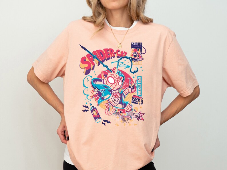 K&ouml;nnte beinhalten: Ein pfirsichfarbenes T-Shirt mit einem grafischen Aufdruck eines Comic-Katers, der einen rot-blauen Spiderman-Anzug tr&auml;gt. Die Grafik enth&auml;lt den Text "SPIDER-CAT" und weitere farbenfrohe Details.
