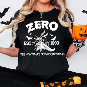May include: A black short-sleeved t-shirt featuring a white graphic of the character Zero from The Nightmare Before Christmas. The design includes the text ZERO, EST. 1993, and THE NIGHTMARE BEFORE CHRISTMAS, along with a spiderweb and bat silhouettes.