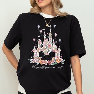 Pu&ograve; includere: Maglietta nera con un design floreale di un castello e le orecchie di Topolino. Il testo "Happiest place on earth" &egrave; stampato sotto il design.
