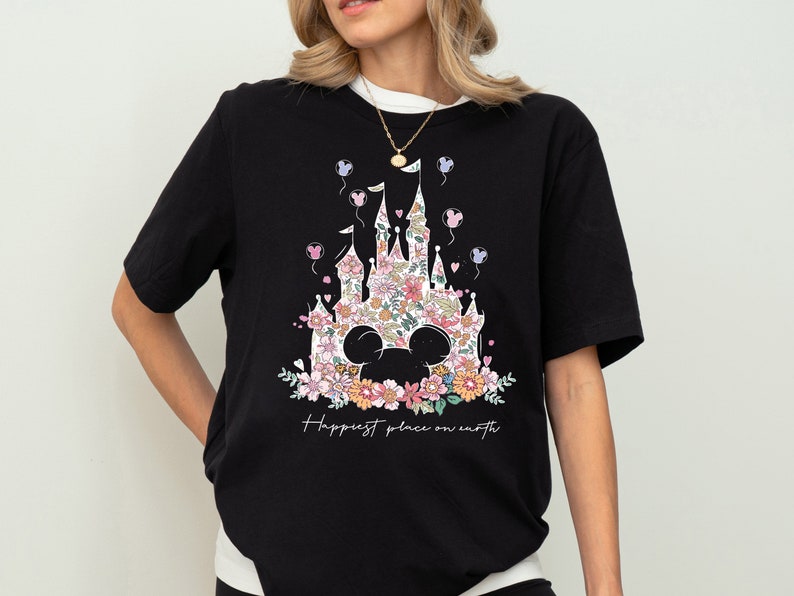 Pu&ograve; includere: Maglietta nera con un design floreale di un castello e le orecchie di Topolino. Il testo "Happiest place on earth" &egrave; stampato sotto il design.
