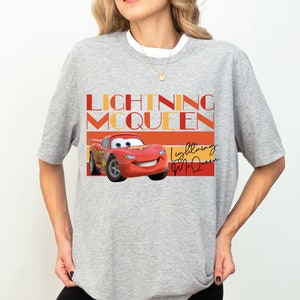 K&ouml;nnte beinhalten: Graues T-Shirt mit einem roten Comic-Rennwagen und dem Text "Lightning McQueen" in Orange und Rot. Das Auto hat ein l&auml;chelndes Gesicht und ist von orangefarbenen und roten Streifen umgeben.