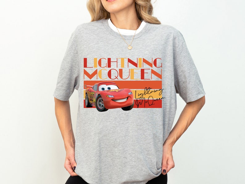 K&ouml;nnte beinhalten: Graues T-Shirt mit einem roten Comic-Rennwagen und dem Text "Lightning McQueen" in Orange und Rot. Das Auto hat ein l&auml;chelndes Gesicht und ist von orangefarbenen und roten Streifen umgeben.