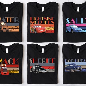 K&ouml;nnte beinhalten: Sechs schwarze T-Shirts mit bunten Retro-Grafiken, die Charaktere aus dem Disney Pixar Film Cars zeigen. Die Shirts zeigen Mater, Lightning McQueen, Sally, Mack, Sheriff und Doc Hudson.