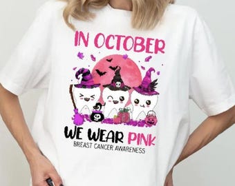 May include: A white t-shirt featuring a graphic of three cartoon teeth wearing purple and black witch hats. The text reads IN OCTOBER WE WEAR PINK BREAST CANCER AWARENESS. The design includes a pink moon, bats, a broom, and a small pumpkin bucket.