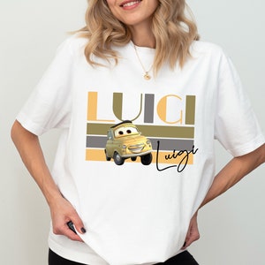 K&ouml;nnte beinhalten: Wei&szlig;es T-Shirt mit einem gelben Comic-Auto und dem Text "Luigi" in braunen, grauen und gelben Streifen.