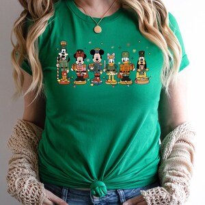 May include: A bright green short-sleeved t-shirt featuring a graphic print of six cartoon characters dressed as nutcracker soldiers. The characters are arranged in a row, each standing on a circular base and connected by a string of colorful holiday lights.