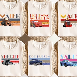 K&ouml;nnte beinhalten: Sechs wei&szlig;e T-Shirts mit verschiedenen Cars-Charakteren bedruckt. Die Charaktere sind Mater, Lightning McQueen, Mack, Sheriff, Doc Hudson und Sally. Jedes Shirt hat ein Retro-Design mit einem bunten Streifenmuster und dem Namen des Charakters in fetten Buchstaben.