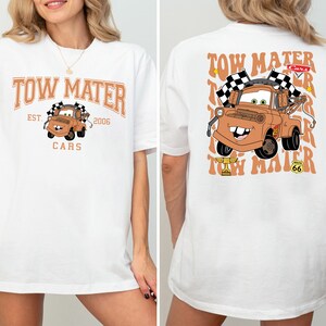 K&ouml;nnte beinhalten: Wei&szlig;es T-Shirt mit einem Cartoon-Bild von Tow Mater aus dem Film Cars. Die Vorderseite des Shirts tr&auml;gt den Schriftzug "Tow Mater" mit "Est. 2006" und "Cars" darunter. Die R&uuml;ckseite des Shirts zeigt ein gr&ouml;&szlig;eres Bild von Tow Mater mit dem Text "Tow Mater", der im Retro-Stil wiederholt wird.