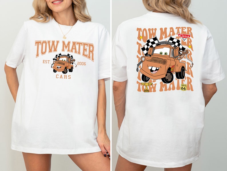 K&ouml;nnte beinhalten: Wei&szlig;es T-Shirt mit einem Cartoon-Bild von Tow Mater aus dem Film Cars. Die Vorderseite des Shirts tr&auml;gt den Schriftzug "Tow Mater" mit "Est. 2006" und "Cars" darunter. Die R&uuml;ckseite des Shirts zeigt ein gr&ouml;&szlig;eres Bild von Tow Mater mit dem Text "Tow Mater", der im Retro-Stil wiederholt wird.