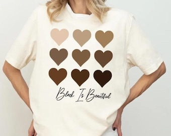 May include: A cream-colored short-sleeve t-shirt featuring a grid of nine hearts in varying shades of brown, arranged in three rows. Below the hearts, the text "Black Is Beautiful" is printed in a black, cursive, handwritten-style font.
