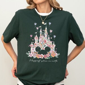 Pu&ograve; includere: Una maglietta verde scuro con una stampa floreale del castello di Cenerentola e il testo "Happiest place on earth".