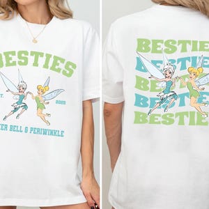 Op de afbeelding: Wit T-shirt met het woord "BESTIES" in groene blokletters en een afbeelding van Tinker Bell en Periwinkle. Het shirt bevat ook de tekst "EST. 2023" en "TINKER BELL & PERIWINKLE" in teal.