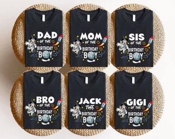 Peut inclure: Six t-shirts noirs sur le thème de l'espace avec un Mickey Mouse astronaute en dessin animé. Les t-shirts portent le texte "Papa du garçon d'anniversaire", "Maman du garçon d'anniversaire", "Sœur du garçon d'anniversaire", "Frère du garçon d'anniversaire", "Jack le garçon d'anniversaire" et "Gigi du garçon d'anniversaire".