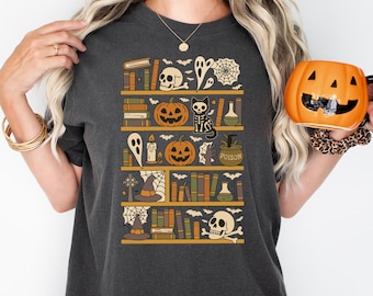May include: A charcoal gray t-shirt featuring a graphic print of bookshelves filled with Halloween-themed items including skulls, pumpkins, a skeleton cat, witch hats, spider webs, ghosts, potion bottles, and various books with bats flying above.