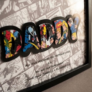 Pode incluir: Obra de arte emoldurada com a palavra "DADDY" em letras grossas, estilo banda desenhada. As letras são preenchidas com ilustrações coloridas de super-heróis e colocadas sobre um fundo de banda desenhada a preto e branco. Inclui uma mensagem personalizada.