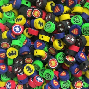 May include: A collection of small, cylindrical, multi-coloured beads featuring various superhero-themed graphic prints, including logos and character masks in red, blue, yellow, green, and black, each with a central hole for threading.