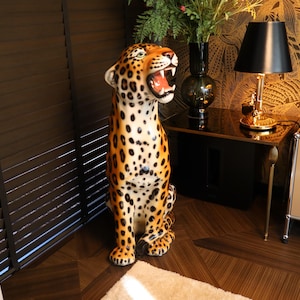 Op de afbeelding: Een decoratief luipaardbeeld met open mond, met oranje, zwarte en witte vlekken. Het beeld staat in de buurt van een donkere houten jaloezie en een bijzettafel met een lamp en een vaas.