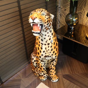 Op de afbeelding: Een keramisch luipaardbeeldje met een gevlekt bruin en zwart patroon. Het beeldje staat op een houten vloer met een zwarte en gouden tafel op de achtergrond.