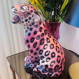 Peut inclure: Une figurine de jaguar en céramique rose et noire, avec une gueule ouverte et des taches noires. La figurine décorative a une finition brillante et est posée sur une table miroir.