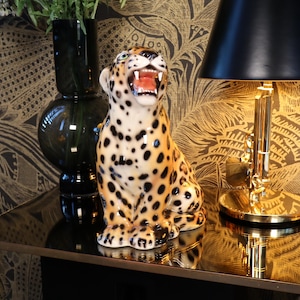 Op de afbeelding: Een keramiek luipaardbeeldje met zwarte en bruine vlekken, dat op een zwarte tafel staat met een gouden lamp met een zwarte lampenkap. De tafel heeft een gouden rand en staat voor een goud-zwart gepatroneerd behang.
