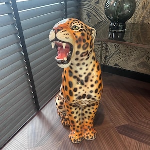 Può includere: Una statuetta di leopardo in ceramica con macchie arancioni e nere, seduta su un pavimento di legno. Il leopardo ha la bocca aperta e guarda a sinistra.