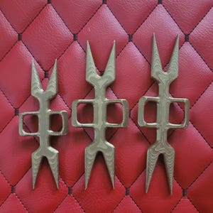 May include: Three gold-colored, abstract metal sculptures with a symmetrical design. Each piece has a central body with a rectangular opening and three pointed prongs at the top and bottom. The sculptures are arranged on a red, quilted surface.