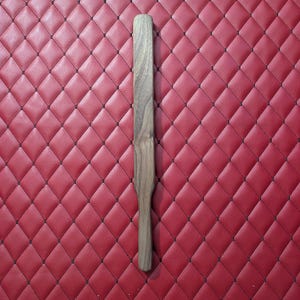 May include: A long, slender wooden paddle with a smooth, natural brown grain finish. The object features a rounded handle that transitions into a wider, flat rectangular blade section, showcasing a consistent wood texture throughout its length.