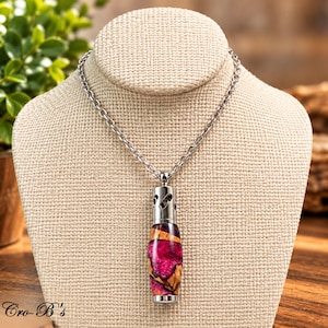 May include: A silver-toned metal pendant necklace featuring a cylindrical diffuser charm. The charm is crafted from a combination of natural wood and vibrant magenta resin with a swirling pattern. The pendant hangs from a simple silver-toned chain.