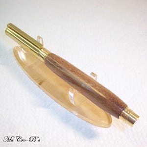 May include: A handcrafted pen with a wooden barrel and gold-colored metal accents. The pen is displayed on a clear acrylic stand. The pen's barrel has a rich, brown wood grain pattern. The pen's metal components include the tip, clip, and end cap.