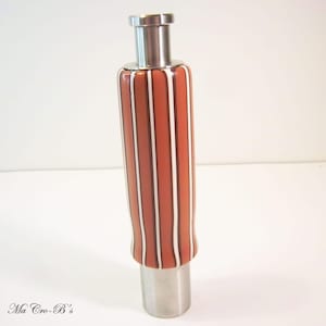 May include: A decorative bottle stopper with a stainless steel top and base. The body features vertical stripes in shades of brown, white, and dark brown. The text "Ma Cro-B's" is visible at the bottom left.