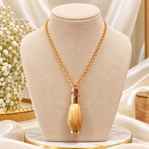May include: A gold-toned chain necklace featuring a pendant shaped like a small, smooth, light brown wooden bottle with a metallic gold cap. The pendant has a cylindrical top section with small cutouts and a rounded, polished wooden base.