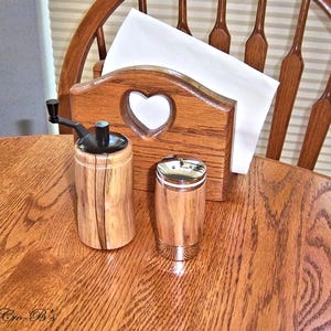 May include: A wooden napkin holder with a heart-shaped cutout, a wooden pepper grinder, and a small, silver-topped container sit on a wooden table. The grinder has a black handle. A wooden chair is in the background.