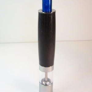 May include: A tall, cylindrical pepper grinder with a black wooden body and a clear, transparent lower section. The top features a vibrant blue accent and a silver-colored metal band. The base is also silver-colored metal.