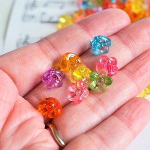 May include: A hand holding a variety of colorful flower-shaped plastic beads. The beads are translucent and have a glossy finish. The beads are in shades of pink, orange, yellow, green, and blue.