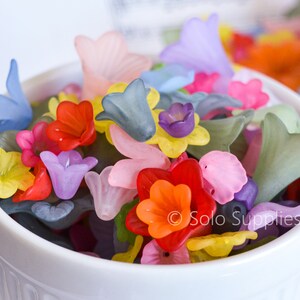 May include: A white bowl filled with colourful flower-shaped beads. The beads are made of translucent plastic and come in a variety of colours, including pink, purple, blue, yellow, orange, and red.