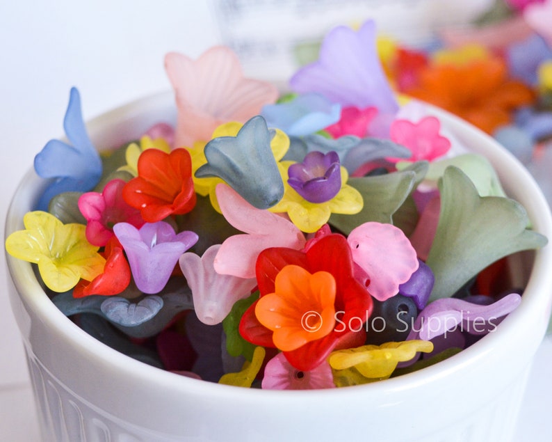 May include: A white bowl filled with colourful flower-shaped beads. The beads are made of translucent plastic and come in a variety of colours, including pink, purple, blue, yellow, orange, and red.