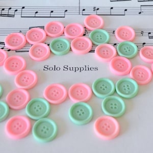 May include: A collection of pink and mint green buttons with four holes, scattered on a surface with musical notes in the background. The text "Solo Supplies" is visible. These buttons are ideal for sewing and craft projects.