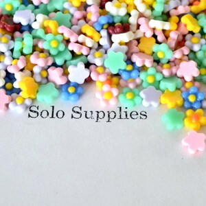 May include: A collection of colorful flower-shaped embellishments in various shades of pink, yellow, blue, green, and white. The text "Solo Supplies" is printed in black on a white background. These small, decorative flowers are likely used for crafts.