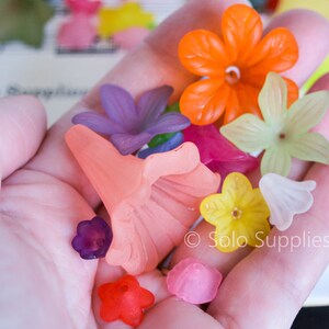 May include: Assortment of colourful plastic flower-shaped beads in various colours, including orange, pink, yellow, purple, and green. The beads are in a hand, and the text "Solo Supplies" is visible in the bottom right corner.