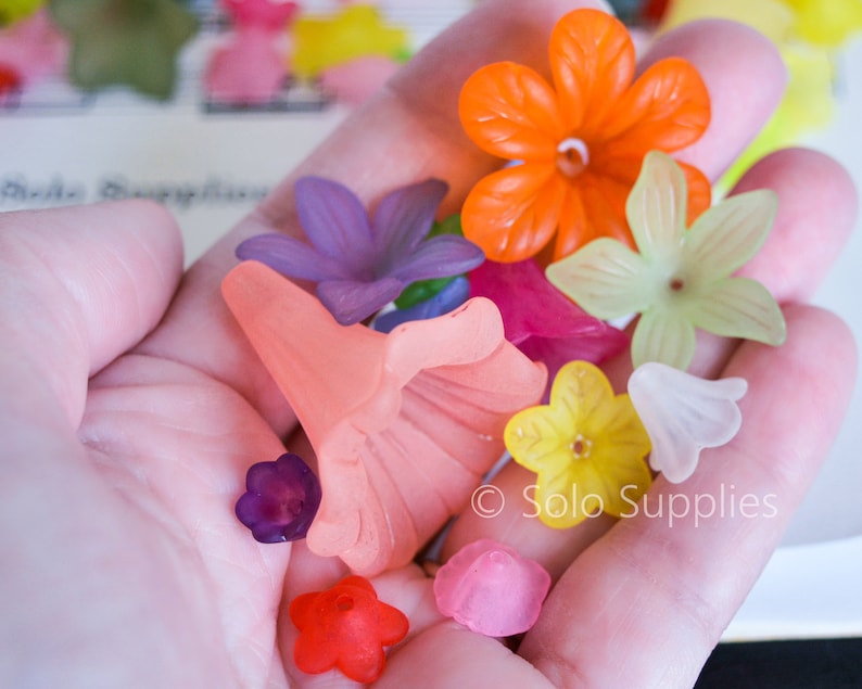 May include: Assortment of colourful plastic flower-shaped beads in various colours, including orange, pink, yellow, purple, and green. The beads are in a hand, and the text "Solo Supplies" is visible in the bottom right corner.