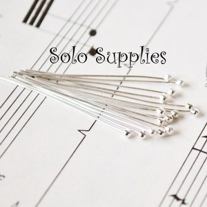 May include: A close-up of a bundle of silver-toned head pins with round heads. The pins are arranged in a fan shape on a sheet of music with the text "Solo Supplies" printed at the top.