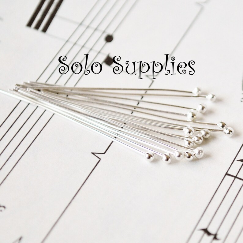 May include: A close-up of a bundle of silver-toned head pins with round heads. The pins are arranged in a fan shape on a sheet of music with the text "Solo Supplies" printed at the top.
