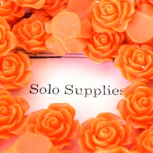 May include: Orange resin rose embellishments arranged around the text "Solo Supplies". The roses are a vibrant orange color and appear to be made of a glossy material. These decorative flowers are ideal for craft projects.