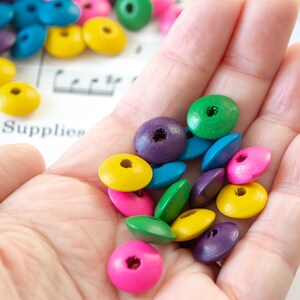 May include: A hand holding a variety of colorful wooden beads. The beads are in shades of green, blue, purple, pink, and yellow. The beads are all round and have a hole in the center.