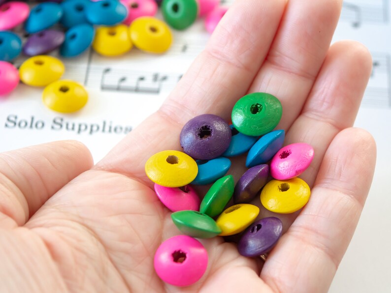 May include: A hand holding a variety of colorful wooden beads. The beads are in shades of green, blue, purple, pink, and yellow. The beads are all round and have a hole in the center.