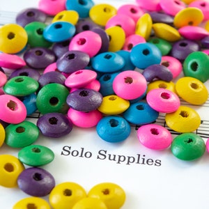 May include: A collection of colorful wooden beads in various shapes and sizes, including circles, squares, and ovals. The beads are scattered on a sheet of music with the text "Solo Supplies".