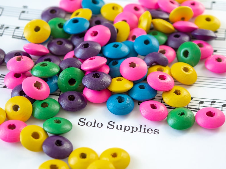May include: A collection of colorful wooden beads in various shapes and sizes, including circles, squares, and ovals. The beads are scattered on a sheet of music with the text "Solo Supplies".