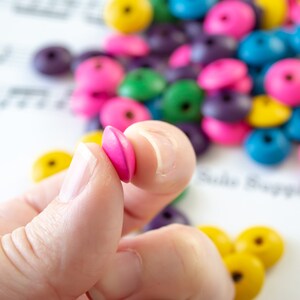 May include: A hand holding two pink wooden beads. The beads are round and have a smooth surface. The beads are in focus, while the background is blurred. The background includes a variety of colorful wooden beads.